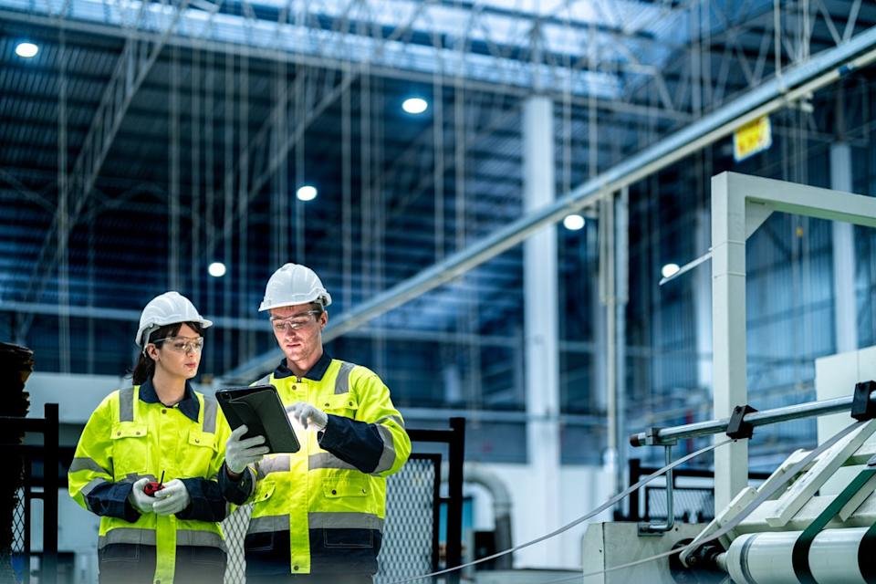 Two engineers in hardhats look at a table while standing in an industrial factory setting.
