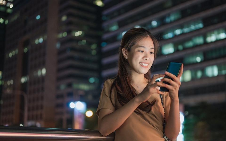 A person smiling, looking at their phone, with a nighttime city skyline in the background.