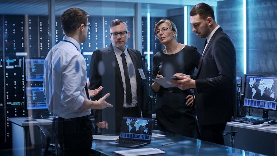 Several people speaking to one another in a dimly lit room with servers in background.
