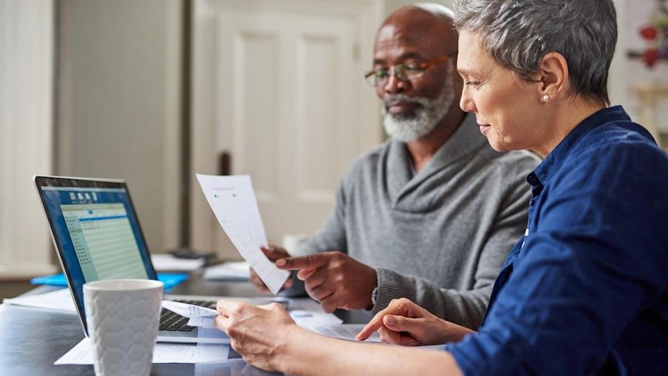Two senior individuals are seated at a table, focused on financial documents and a silver laptop. An African American man with a gray beard and glasses holds a paper, pointing at it. Next to him, a Caucasian woman with short gray hair in a blue shirt holds other papers and points at something on the table. A white textured coffee mug is in the foreground.