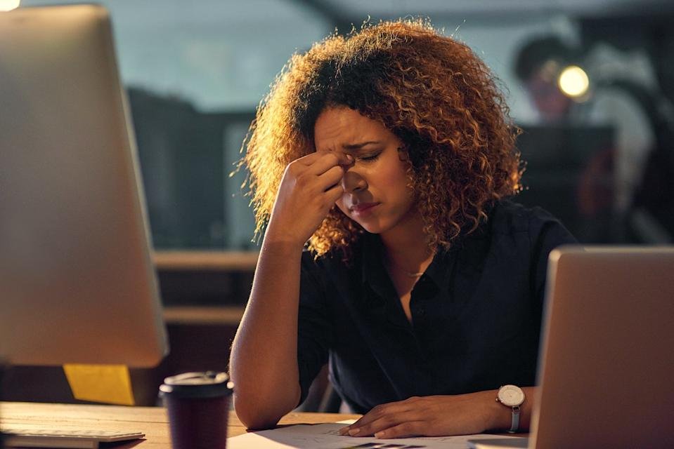 A person sits a desk in front of a computer late at night, touching the bridge of their nose in frustration.