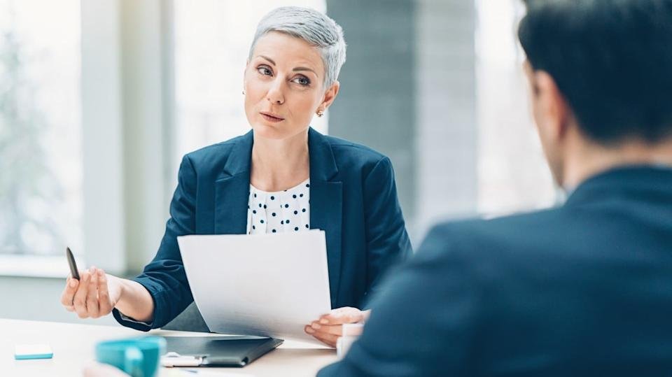 A woman with short grey hair, wearing a navy blazer and polka-dot top, sits at a table holding a pen and documents, looking up while speaking. Across from her, a man is partially visible from behind, listening intently in a bright, modern office setting.