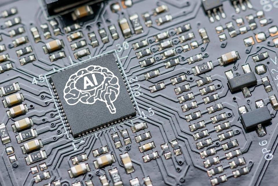 A close-up shot of a dark grey circuit board with a prominent black square microchip. The chip features a white outline of a human brain with the letters 'AI' inside it. Surrounding the chip are many small silver and beige electronic components connected by etched pathways.